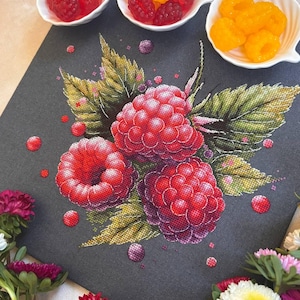 Puede incluir: Un diseño de punto de cruz con tres grandes frambuesas rojas con hojas verdes y amarillas sobre un fondo gris oscuro. Pequeñas cuentas rojas y moradas están esparcidas alrededor de las frambuesas. Tres cuencos blancos pequeños contienen frambuesas frescas.