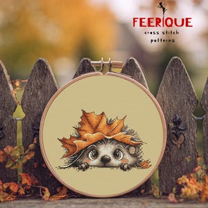 May include: A cross-stitch pattern featuring a hedgehog peeking out from under a large orange autumn leaf. The design is set in a wooden embroidery hoop. The text "FEERIQUE cross stitch patterns" is displayed in red.