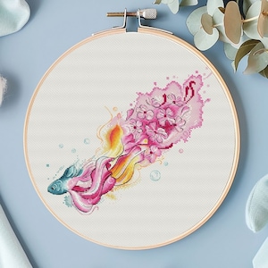 May include: A cross-stitch pattern featuring a blue and pink betta fish with a pink and yellow tail, surrounded by pink flowers and bubbles.