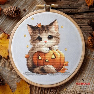 May include: A cross stitch pattern featuring a cute cartoon cat holding a jack-o'-lantern pumpkin. The cat is brown and white with big blue eyes. The pumpkin is orange with a carved face. The background is a light gray with a few yellow stars.