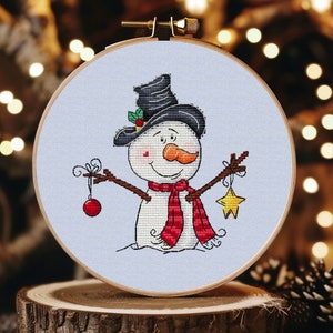May include: A cross-stitch embroidery of a snowman wearing a black top hat, red scarf, and holding a red bauble and a gold star. The snowman is on a light blue background.