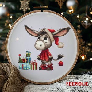 May include: A cross stitch pattern featuring a cute gray donkey wearing a red Santa hat and holding a red ornament. The donkey is standing in front of a pile of wrapped Christmas presents.