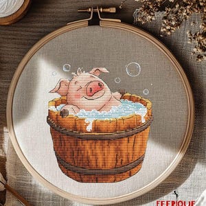 May include: Cross stitch pattern of a pink pig taking a bath in a wooden bucket with bubbles. The pig is smiling and has its eyes closed. The bucket is brown and has a white rim. The background is a light brown fabric.