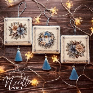 May include: Three white square Christmas decorations with blue tassels. Each decoration features a different embroidered design: a floral bouquet, a wreath, and a floral arrangement. The decorations are hanging from a string of white lights.