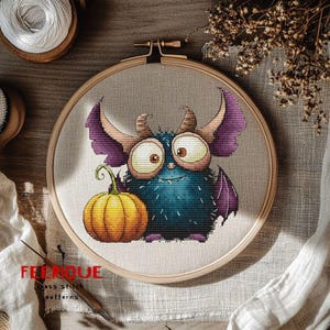 Peut inclure: Broderie au point de croix d'un monstre de dessin anim&eacute; avec de grands yeux, des cornes et des ailes violettes, tenant une citrouille. Le motif est dans un cercle &agrave; broder en bois. Le texte "FELIQUE cross stitch patterns" est visible.