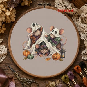 May include: A cross stitch pattern in a wooden hoop featuring three mice wearing white ghost costumes. One mouse wears a purple witch hat. The design includes two orange pumpkins, a green pumpkin, and a small lantern on a light gray fabric background.