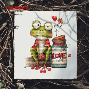 Puede incluir: Patrón de punto de cruz que muestra una rana verde con una camisa roja, sentada junto a un tarro con la etiqueta "LOVE POTION". La rana tiene ojos grandes y pies rojos. Una flor y un corazón están encima del tarro.