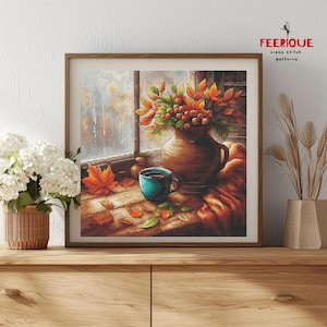 May include: Cross-stitch pattern of a cozy autumn scene. A brown vase holds fall foliage with red berries. A teal mug sits on a wooden surface with scattered leaves. The framed artwork is displayed on a wooden cabinet.