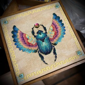 May include: A framed cross-stitch embroidery depicting a colourful scarab beetle with iridescent wings. The beetle is holding a pink gem in its front legs.
