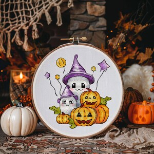 May include: A white embroidery hoop with a Halloween-themed cross-stitch design. The design features three ghosts, two with hats, and three pumpkins. The ghosts have smiling faces and the pumpkins have jack-o'-lantern faces.