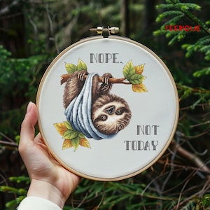 May include: A cross-stitch hoop with a sloth design and the text "Nope, Not Today." The sloth is brown and tan, with a blue blanket. The leaves are green and yellow. The hoop is held in a hand. The background is blurred.