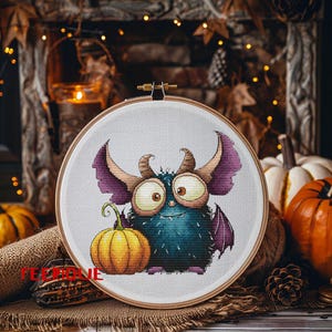Peut inclure: Un cercle &agrave; broder au point de croix repr&eacute;sentant un monstre de dessin anim&eacute; avec de grands yeux, des cornes et des ailes, tenant une petite citrouille orange. Le monstre est principalement bleu avec des accents violets. Fond rustique d'automne.