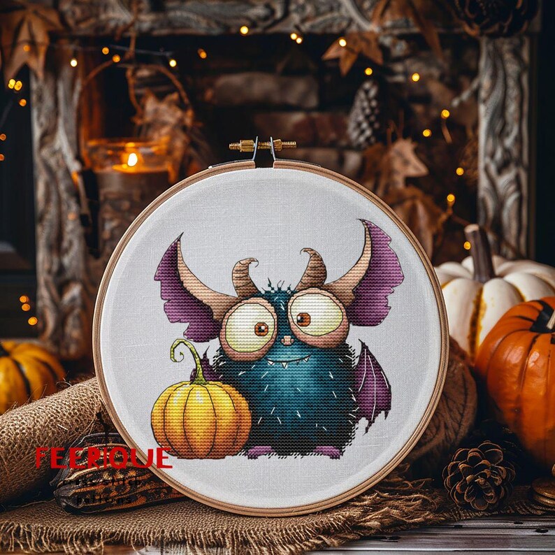 Peut inclure: Un cercle &agrave; broder au point de croix repr&eacute;sentant un monstre de dessin anim&eacute; avec de grands yeux, des cornes et des ailes, tenant une petite citrouille orange. Le monstre est principalement bleu avec des accents violets. Fond rustique d'automne.