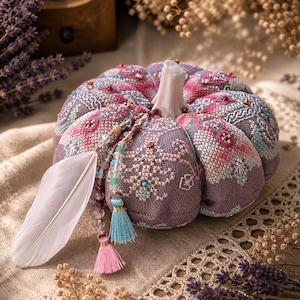 Può includere: Una zucca decorativa in tessuto con intricati ricami a punto croce nelle tonalità del malva, rosa e blu. La zucca è sormontata da un gambo bianco e decorata con un ciondolo composto da perline trasparenti, una piuma bianca e due piccole nappe in azzurro e rosa.