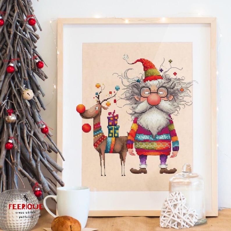 May include: A cross stitch pattern featuring a whimsical Santa Claus with a long white beard and glasses, wearing a colorful sweater and a red hat. He is standing next to a reindeer with a red nose and colorful decorations. The pattern is framed and displayed on a table with a Christmas tree in the background.