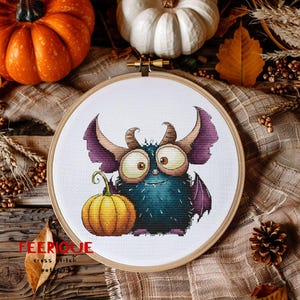 Peut inclure: Une broderie au point de croix dans un cercle en bois repr&eacute;sente un monstre de dessin anim&eacute; avec de grands yeux, des cornes et des ailes, tenant une citrouille. Le monstre est bleu sarcelle et violet. Le texte "FEERIQUE cross stitch patterns" est en bas.