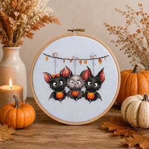 May include: A circular wooden embroidery hoop containing a cross-stitch pattern of three small, fluffy creatures hanging from a string decorated with orange and yellow lights. Each creature holds a small orange pumpkin with a carved face.