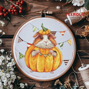 May include: Cross-stitch embroidery of a guinea pig dressed as a pumpkin, with a pumpkin hat and holding a banner. The design is set in a wooden embroidery hoop. Colorful bunting flags are in the background. The text "FEERIQUE cross stitch patterns" is visible.