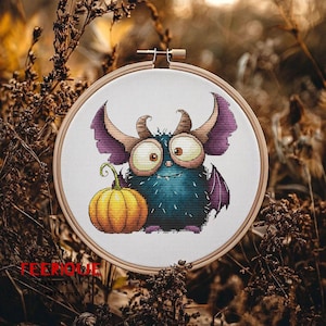 Peut inclure: Broderie au point de croix dans un cercle, repr&eacute;sentant un monstre fantaisiste avec de grands yeux, des cornes et des ailes violettes, tenant une citrouille. Le monstre est principalement bleu avec des accents marron et violets. Le texte "FEERIQUE" est visible.