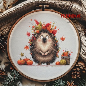 Peut inclure: Un cercle de broderie au point de croix représentant un hérisson souriant orné de feuilles et de baies d'automne. Le motif comprend des citrouilles et le texte "FLEUNIQUE cross stitch patterns". Le cercle est posé sur un fond texturé.