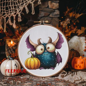 Peut inclure: Un cercle de broderie au point de croix avec un monstre de dessin anim&eacute;, une citrouille jaune et le texte "FEERIQUE CROSS". Le monstre a de grands yeux, des cornes et des ailes violettes. Th&egrave;me d'Halloween.
