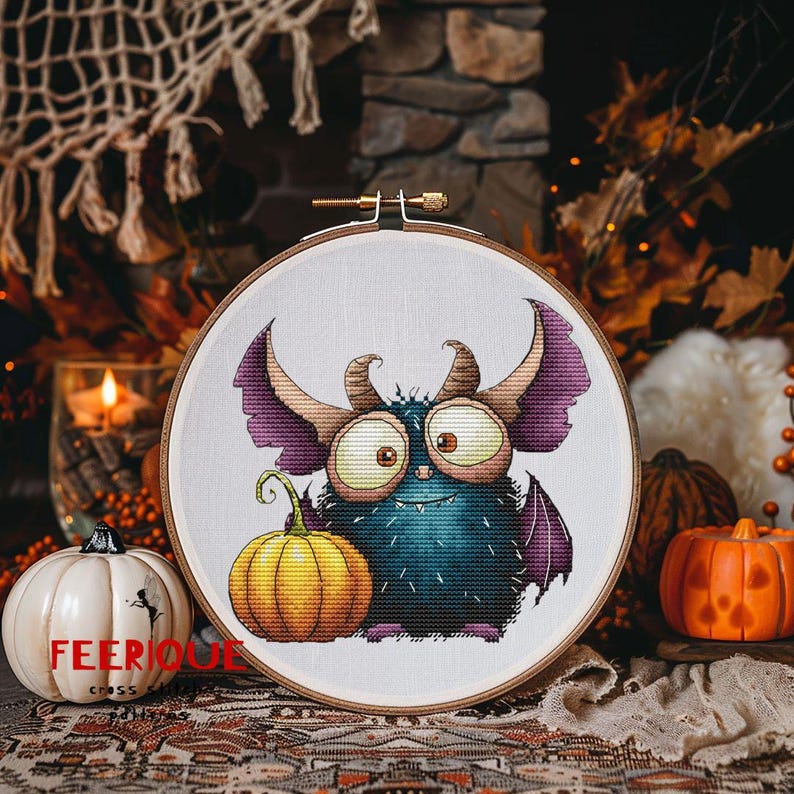 Peut inclure: Un cercle de broderie au point de croix avec un monstre de dessin anim&eacute;, une citrouille jaune et le texte "FEERIQUE CROSS". Le monstre a de grands yeux, des cornes et des ailes violettes. Th&egrave;me d'Halloween.