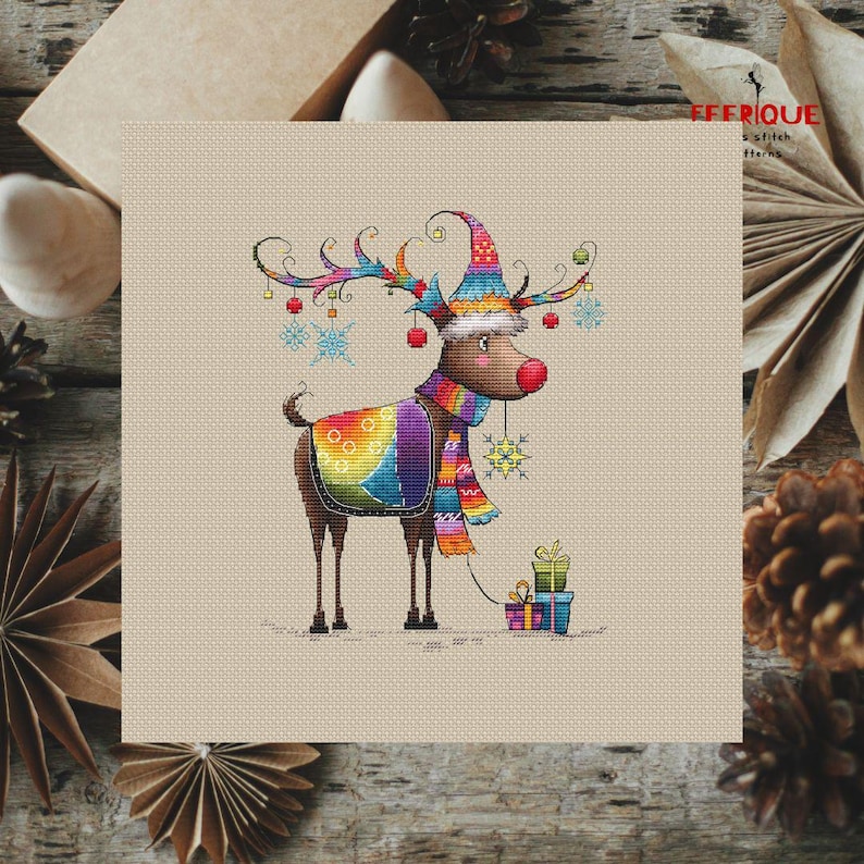 May include: A colorful cross-stitch pattern of a reindeer wearing a rainbow-colored scarf and hat, holding a gift box. The reindeer is surrounded by snowflakes and red ornaments.