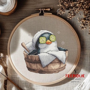 May include: Cross-stitch embroidery of a penguin relaxing in a wooden tub, wearing cucumber eye masks. The penguin is wrapped in a white towel, with bubbles and a wooden spoon. The embroidery is in a wooden hoop.