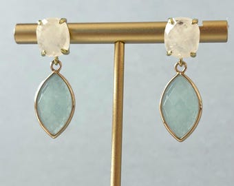 May include: A pair of gold-toned earrings. Each earring features a square, light-colored stone at the top and a teardrop-shaped, pale blue stone pendant. The earrings are displayed on a gold jewelry stand.