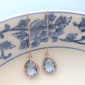 May include: A pair of rose gold drop earrings with teardrop-shaped, light blue gemstones. The earrings hang from thin, curved wires. The gemstones are set in a rose gold bezel. The earrings are displayed on a white plate with a blue floral design.