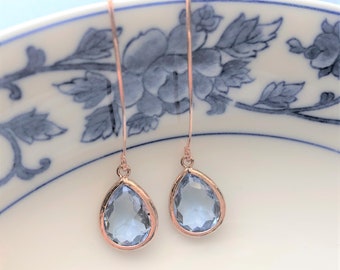 May include: A pair of rose gold drop earrings with teardrop-shaped, light blue gemstones. The earrings hang from thin, curved wires. The gemstones are set in a rose gold bezel. The earrings are displayed on a white plate with a blue floral design.