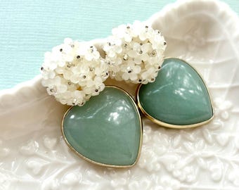 May include: Two heart-shaped, pale green earrings with gold-colored trim rest on a white, floral-patterned dish. Above them are two clusters of small, white, beaded flowers. The background is a light blue.