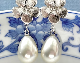 May include: A pair of silver-toned flower earrings with pear-shaped, off-white faux pearls. The earrings have a floral design at the top and dangle below. The earrings are displayed on a white and blue patterned surface.