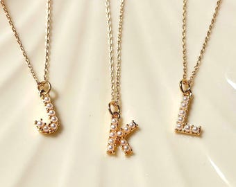 May include: Three gold-toned necklaces featuring letter pendants J, K, and L. Each letter is embellished with small, round, white faux pearls set along the surface of the metal. The necklaces hang from delicate gold-toned link chains.