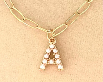 May include: A gold-toned necklace featuring a paperclip chain and a single pendant shaped like the letter A. The letter A is embellished with small, round, white faux pearls set into the metal frame, which hangs from a small circular jump ring.