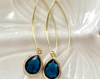 May include: A pair of gold-colored drop earrings. Each earring features a teardrop-shaped, deep blue gemstone set in a gold-colored bezel. The earrings have a long, curved hook design.