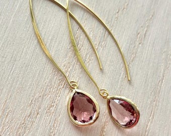 May include: A pair of gold-colored drop earrings. Each earring features a teardrop-shaped, pink gemstone set in a gold-colored bezel. The earrings have a long, curved hook design.