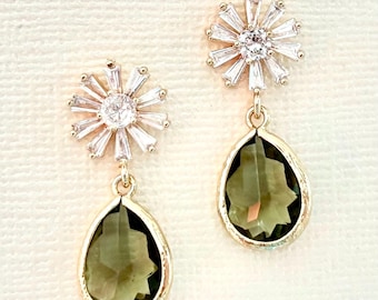 May include: A pair of gold-toned earrings. Each earring features a sparkling, starburst-shaped top with a central clear stone. A teardrop-shaped, olive-green stone hangs below, framed by a gold-colored setting. The earrings are displayed against a cream-colored background.