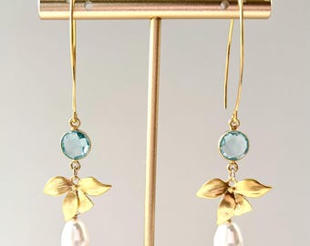 May include: Gold-tone earrings with a light blue gemstone, a gold-colored floral design, and a white teardrop-shaped pearl. The earrings are displayed on a gold-colored stand.