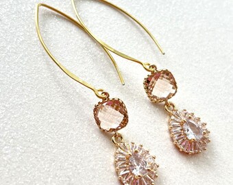 May include: A pair of gold-toned drop earrings. Each earring features a square, light pink gemstone at the top, with a teardrop-shaped cluster of clear and pink crystals below. The earrings have a simple, curved hook design.
