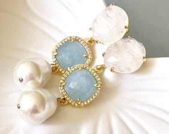 May include: Elegant gold-toned earrings featuring a combination of light blue square-cut stones, white pear-shaped stones, and large white pearl-like drops. The square stones are surrounded by a halo of small, clear crystals. The earrings are displayed on a white, scalloped surface.