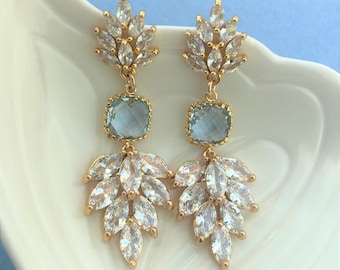 May include: A pair of gold dangle earrings with light blue square-shaped stones and clear crystal accents. The earrings feature a cluster of crystal leaves.