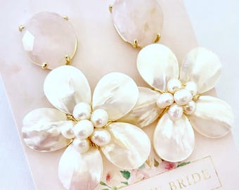 May include: A pair of floral earrings with a light pink gemstone top and white petal-shaped accents. The center of each flower features small, round pearls. The earrings are on a pink card with the text "FROM THE BRIDE".
