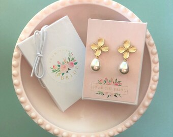 May include: Gold-tone flower earrings with pear-shaped pearl drops displayed on a pink card. The card reads "FROM THE BRIDE" with floral accents. A white gift box with a floral sticker and the text "FROM THE BRIDE" is also present, all set in a pink dish.