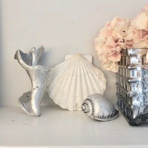May include: A white seashell with a pearl-like finish, a silver seashell with a swirl design, and a silver seashell with a spiral design. The seashells are on a white surface.