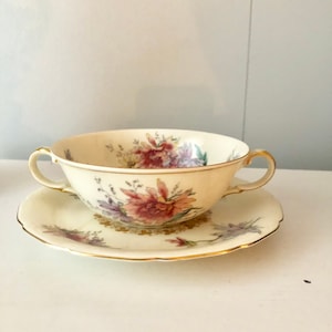 May include: A white porcelain soup bowl with a floral pattern in shades of pink, purple, and green. The bowl has two handles and is sitting on a matching saucer with a gold rim.