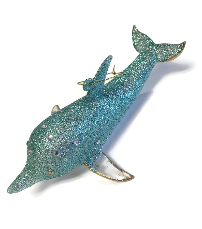 K&ouml;nnte beinhalten: Ein tealblauer Glas-Delfin-Anh&auml;nger mit Glitzer und silbernen Akzenten. Der Delfin hat eine goldene Schnur zum Aufh&auml;ngen.