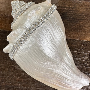 May include: A large, off-white conch shell decorated with multiple strands of clear, sparkling rhinestones wrapped around its spiral body. The shell features a natural, textured surface with a pearlescent sheen and a pointed apex at the top.