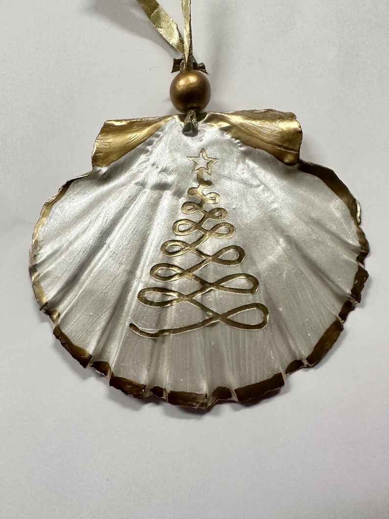 May include: A white seashell ornament with a gold trim and a gold painted Christmas tree design.