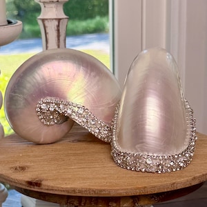Op de afbeelding: Twee iriserende schelpvormige decoratieve objecten met kristallen versieringen. Eén heeft de vorm van een nautilus, de andere van een conch. Beide staan op een ronde, houten sokkel met een witte, sierlijke basis. De schelpen hebben een parelmoerglans.